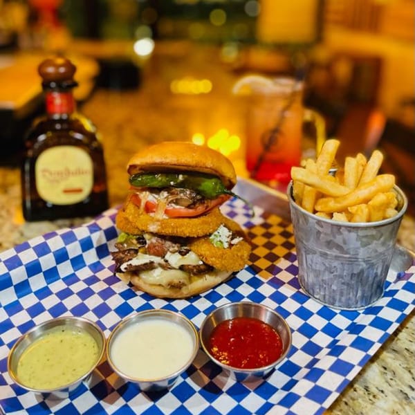 A large burger with onion rings, fries in a bucket, and three dipping sauces on a checkered tray.