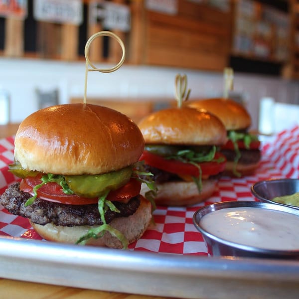 Three Burgar sliders with lettuce, tomato, and pickles on a red and white checkered paper.